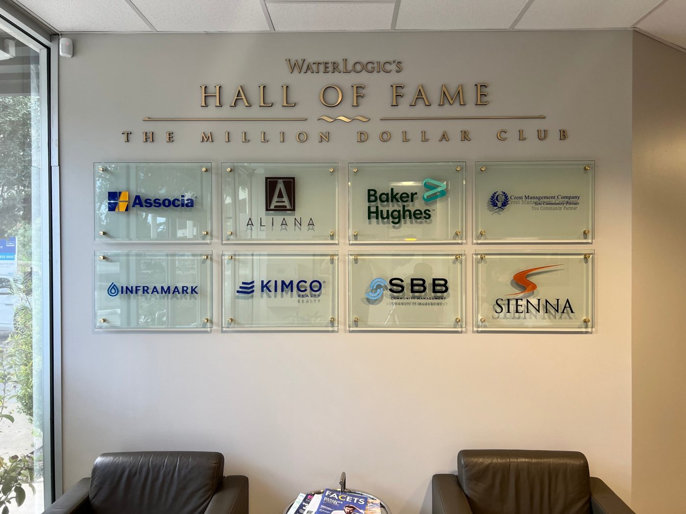 Eight glass plaques carrying company logos, mounted on standoffs in two rows beneath a Hall of Fame wall sign.