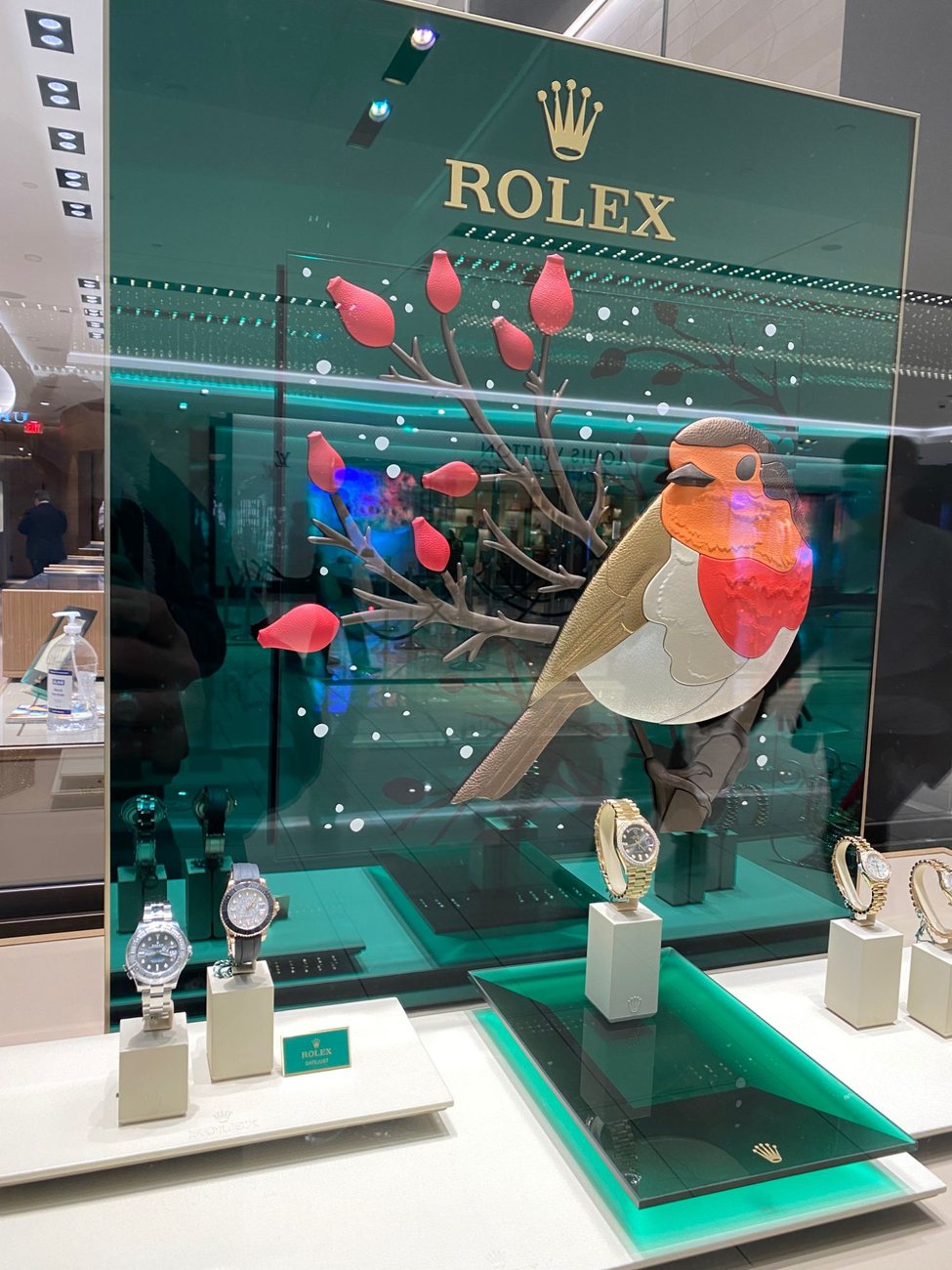 A carved and painted glass display panel showing a robin on a rose-hip branch, standing in a watch boutique window.