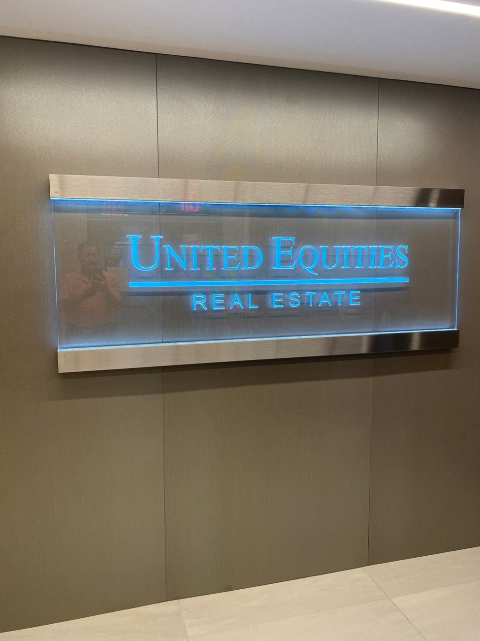 An edge-lit glass lobby sign reading United Equities Real Estate, glowing blue in a brushed metal frame.