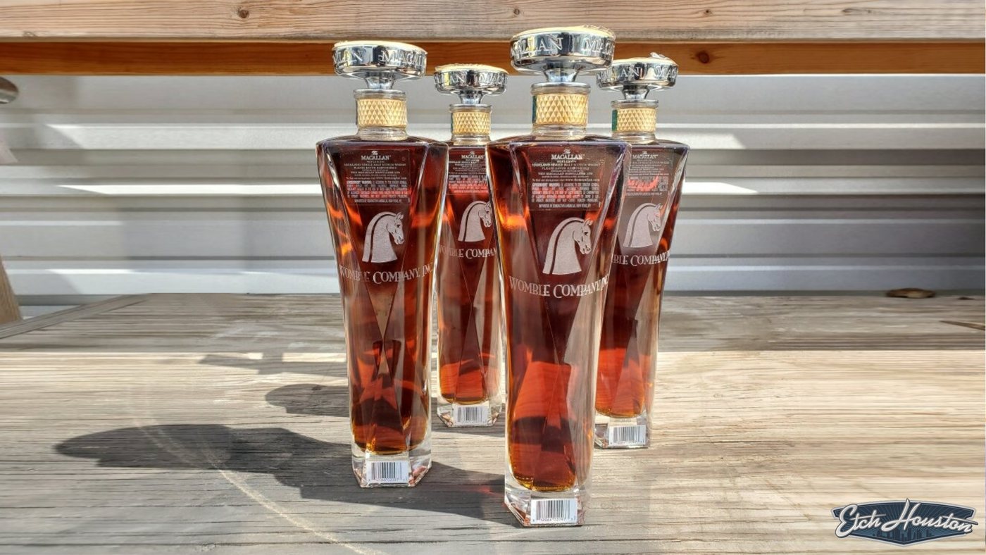 Four Macallan decanters etched with a horse's head and a company name, lit by sunlight on a timber bench.