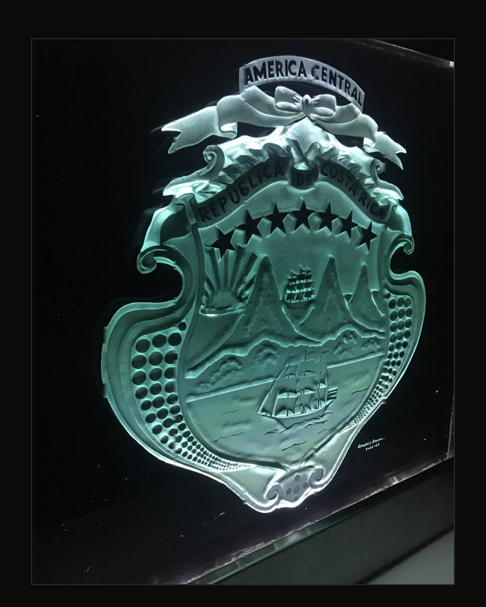 A close view of a national coat of arms sand-carved into dark glass at several depths, the volcanoes, ships and stars standing in frosted relief and lit green from the edge.