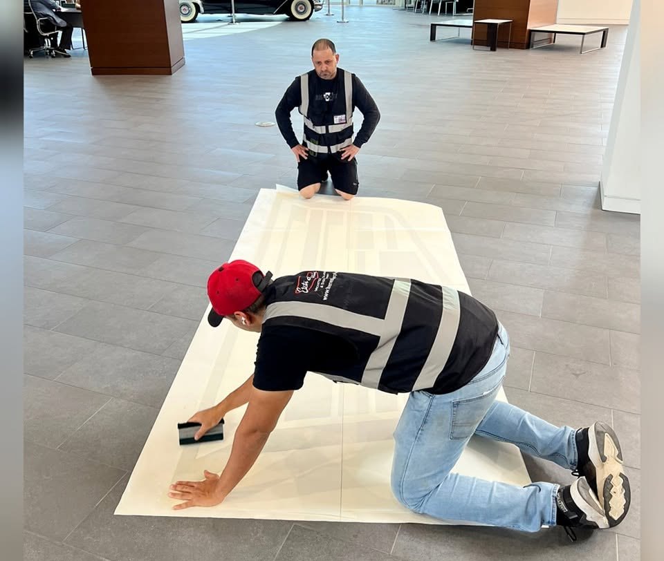 Two installers kneeling on a showroom floor, squeegeeing a large sheet of cut film flat.