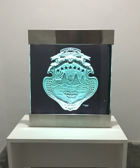 A framed glass artwork of a national crest with ships and volcanoes, sand-carved and lit green from the edge.