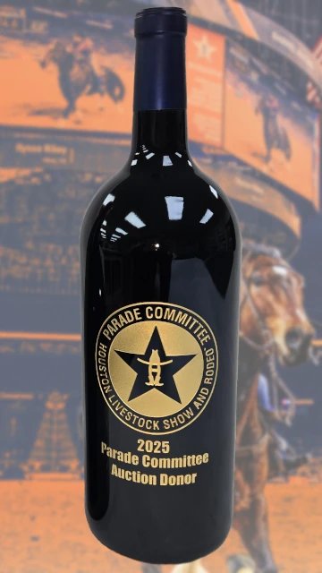A black bottle etched with the 2025 Parade Committee Auction Donor medallion in gold, photographed against a rodeo backdrop.