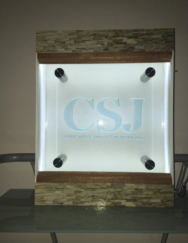 A backlit glass panel carrying the CSJ monogram, mounted on standoffs against a stone wall.