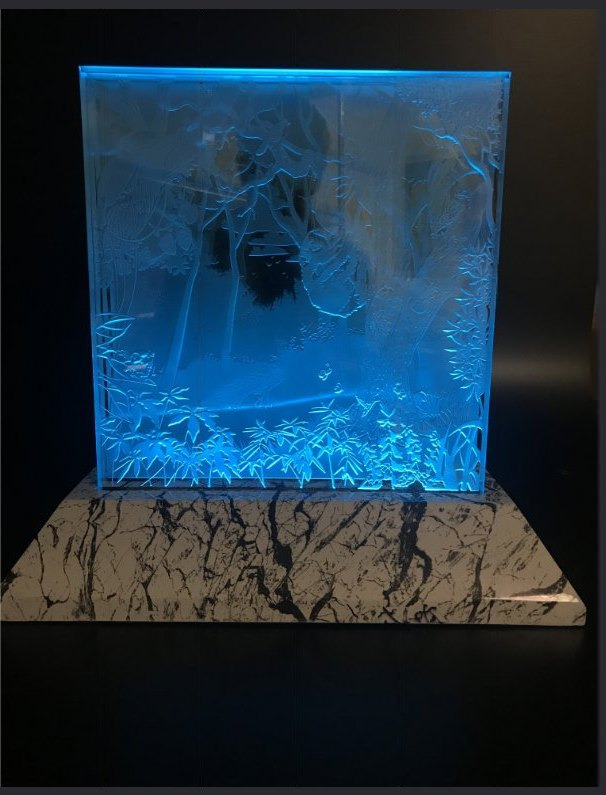 A carved glass panel of a forest scene glowing blue from edge lighting inside a dark display case.