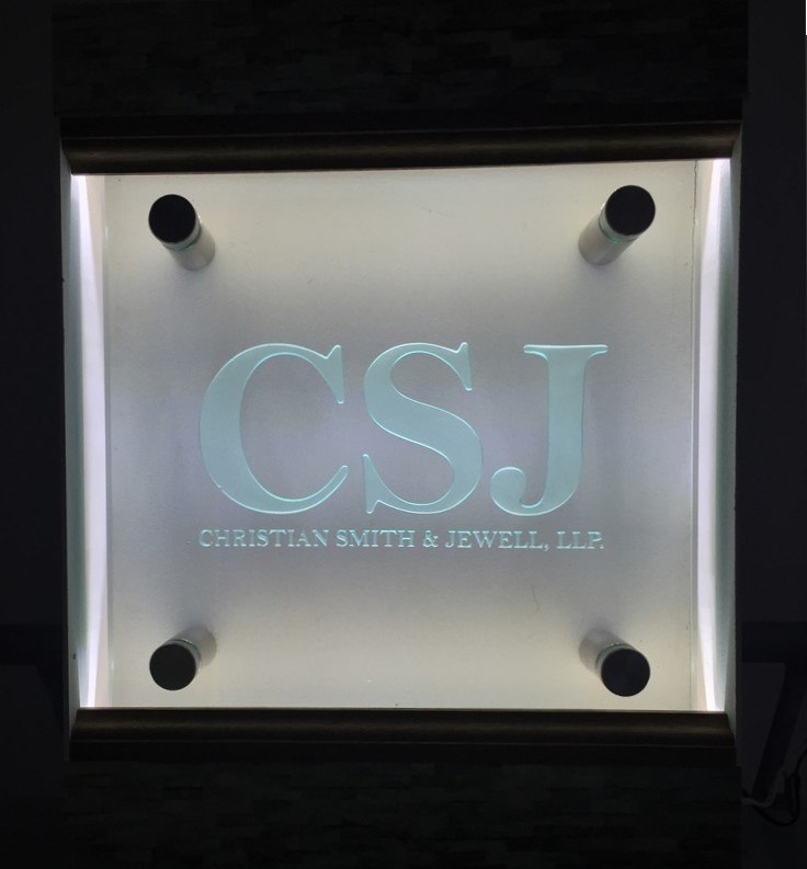 A backlit glass panel etched with the CSJ monogram and Christian Smith and Jewell LLP, mounted on standoffs against a dark wall.