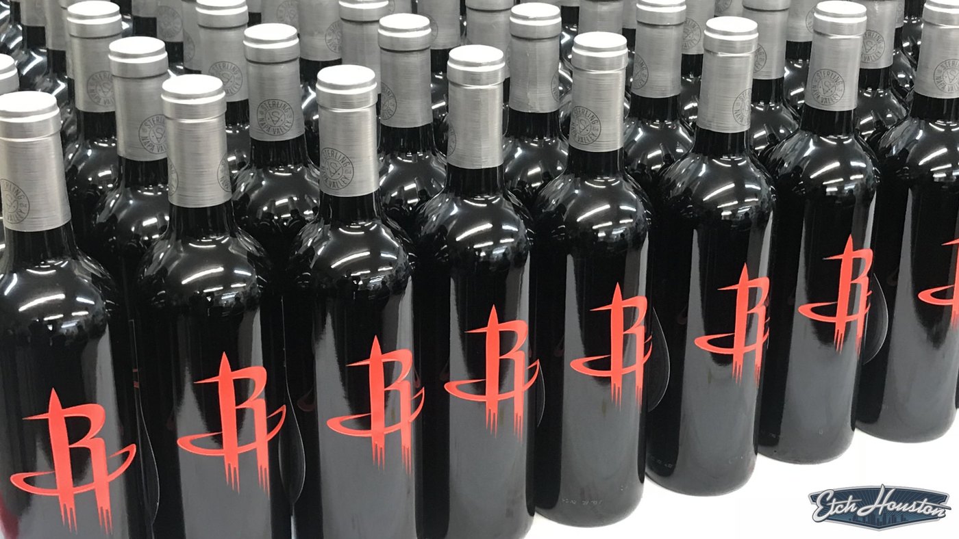 Dozens of black wine bottles arranged in rows, each etched with a red basketball team logo.