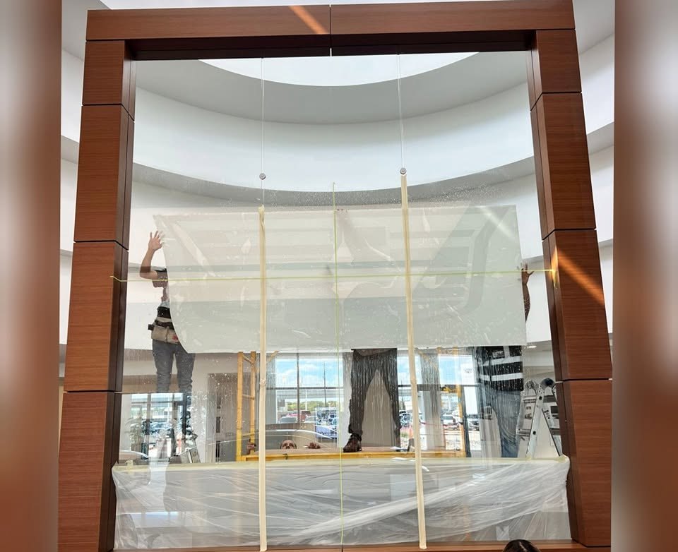Installers holding a full-size cut stencil up against a glass showroom partition to position the marque crest before it is applied.