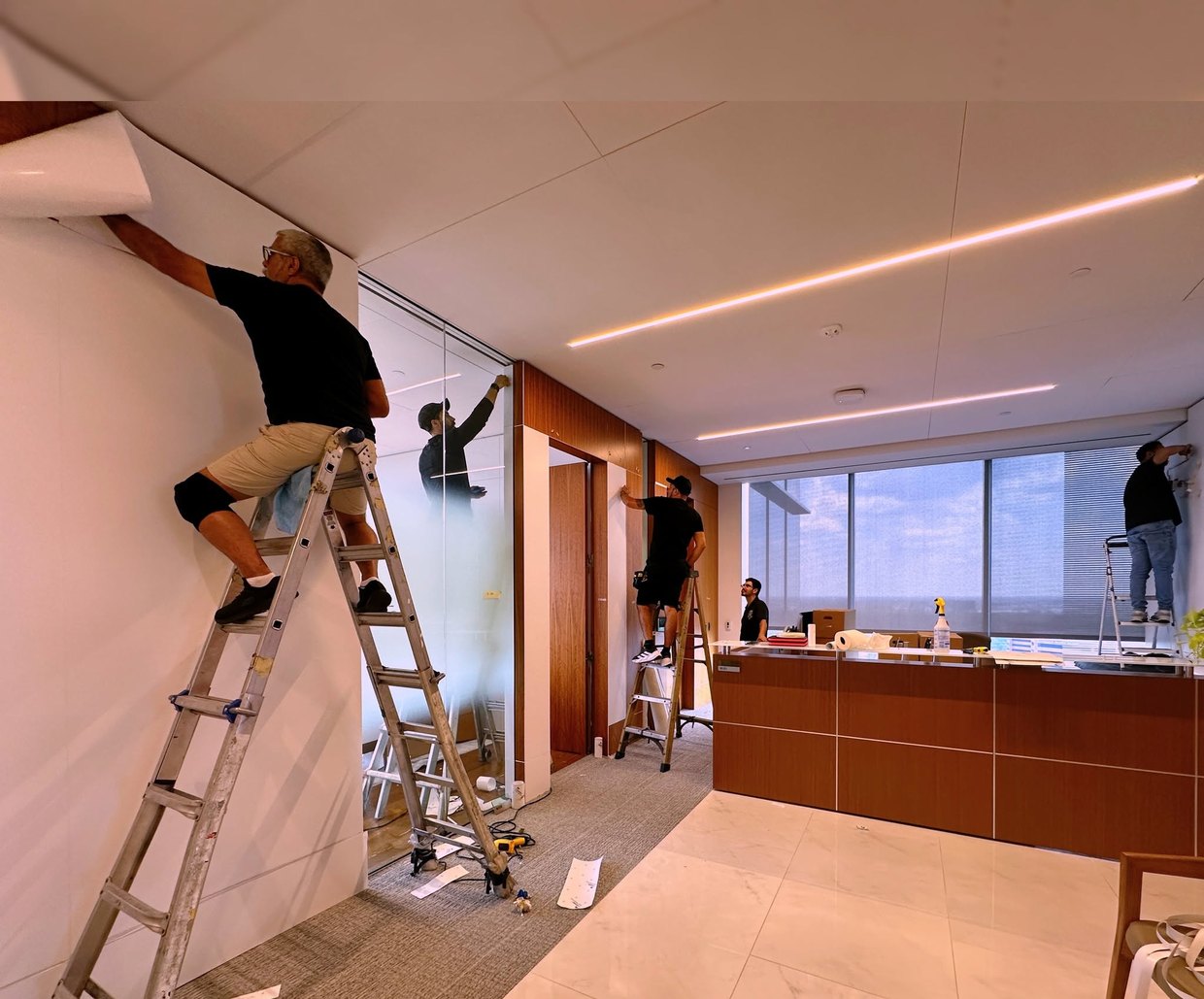 A four-man crew on ladders applying frosted film to a glass office wall in a finished corporate lobby.