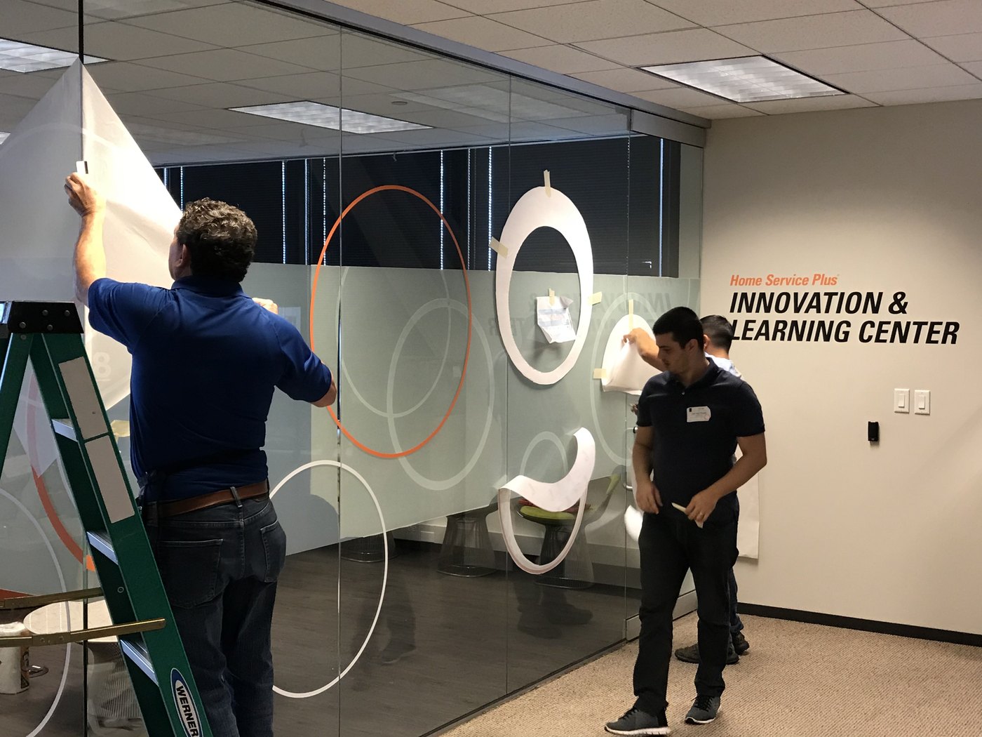 An installer on a ladder laying a large frosted graphic onto office glass, the circular pattern taped into position across the mullions before it is fixed down.