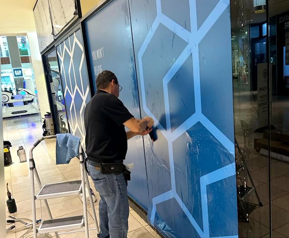 An installer squeegeeing a blue geometric privacy film onto a full-height storefront glass wall inside a shopping centre.
