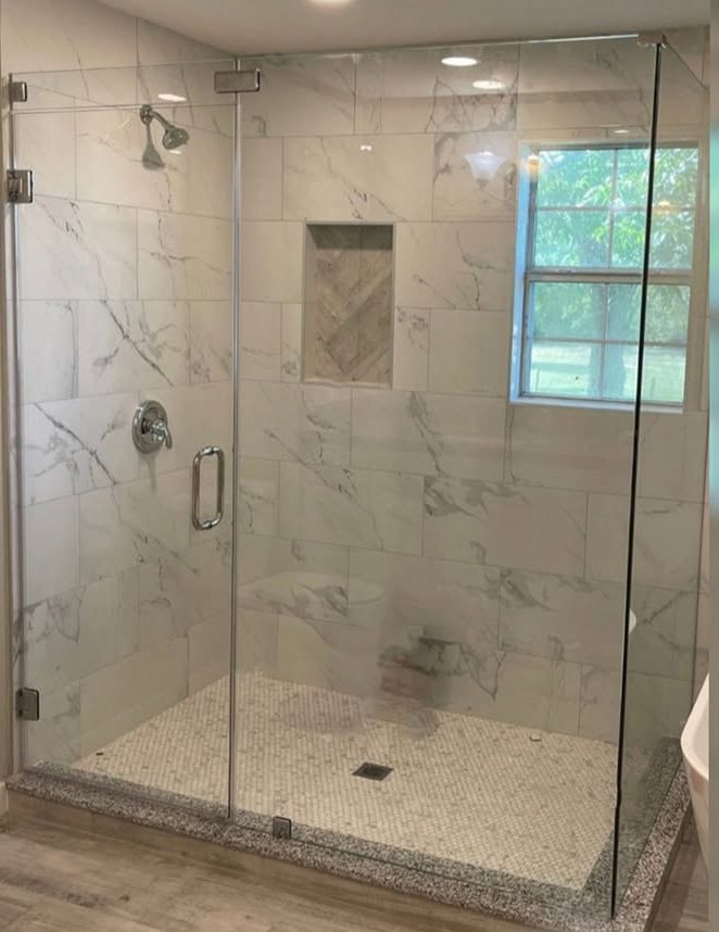 A frameless glass shower enclosure with polished chrome hinges, set against marble tile and a pebble floor.