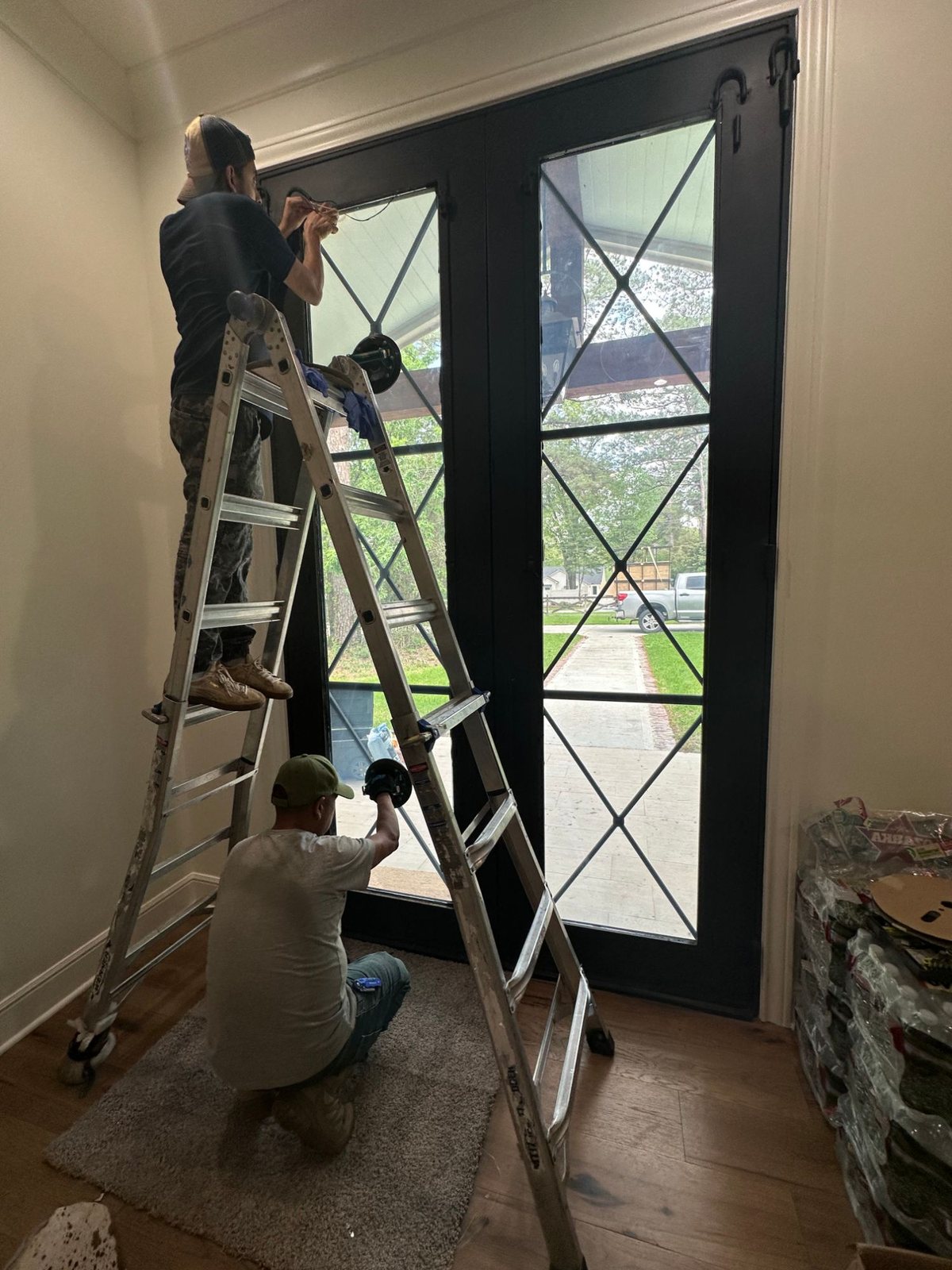 Two installers, one on a ladder, applying switchable film to the tall glass panels of a black steel entry door.
