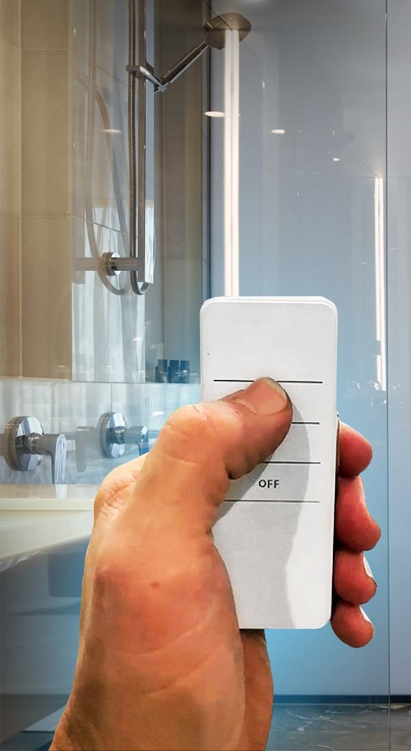 A hand holding a small white remote in front of a frosted glass shower screen.