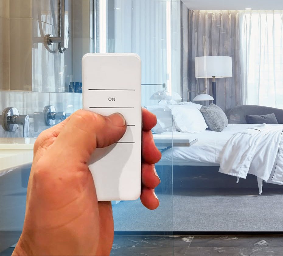 A hand holding the Smart Glass remote, thumb over the ON button, in front of a glass wall that is switched clear.