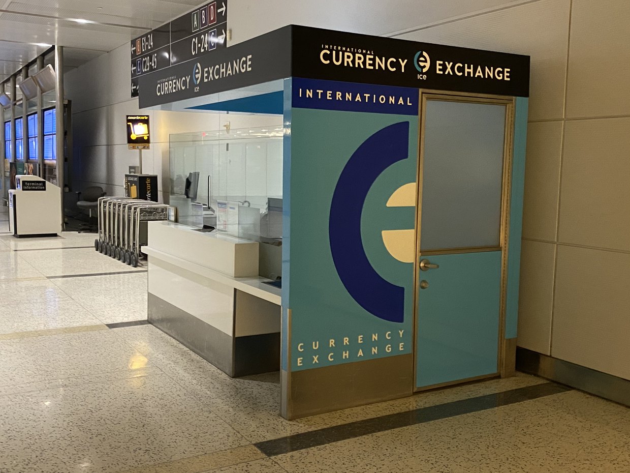 An airport currency exchange kiosk before the work, clad in faded teal panels with a plain door and no glass graphics.